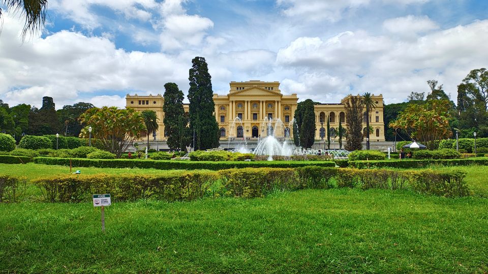 Foto do Museu Paulista, carinhosamente chamado de Museu do Ipiranga é uma imponente edificação de cor bege amarelado com formas clássicas. A sua frente o bonito jardim com a fonte de água.